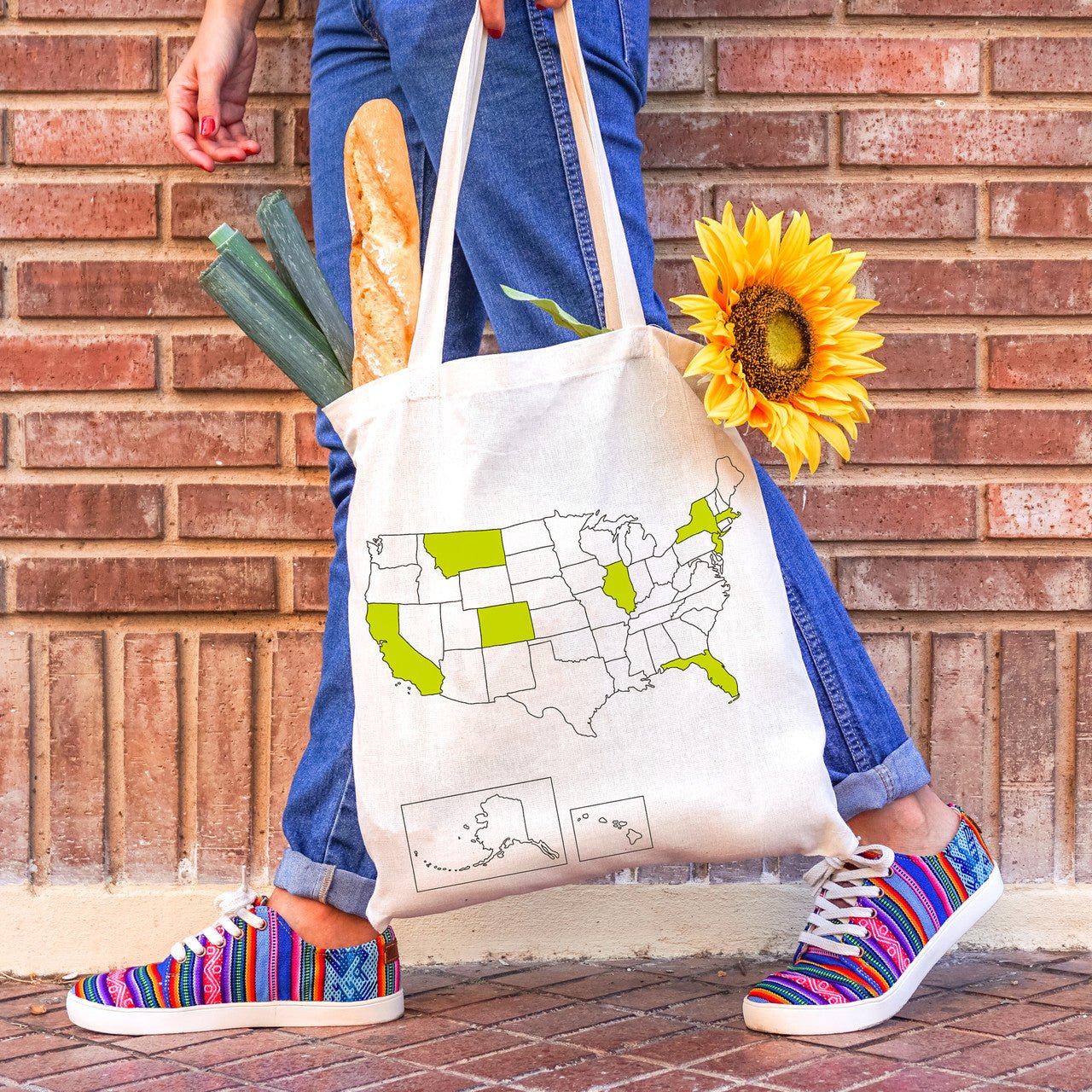 A tote bag featuring a USA map with a few states colored in, filled with leeks, bread and flowers, carried by a woman with colorful shoes on the street
