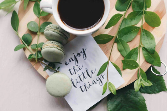 Cup of coffee with green macarons and a note saying 'enjoy the little things' on a wooden board with leaves.