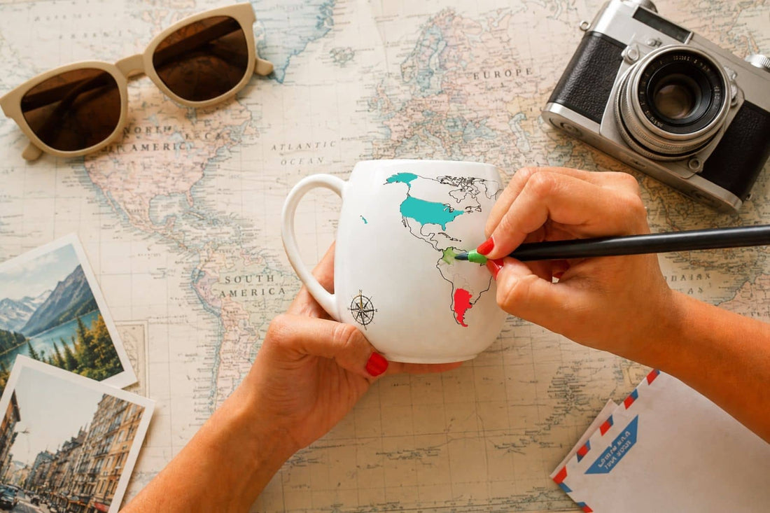 Hands colouring in a country on the world map colour-in mug using a green ceramic pen with travel props in the background.