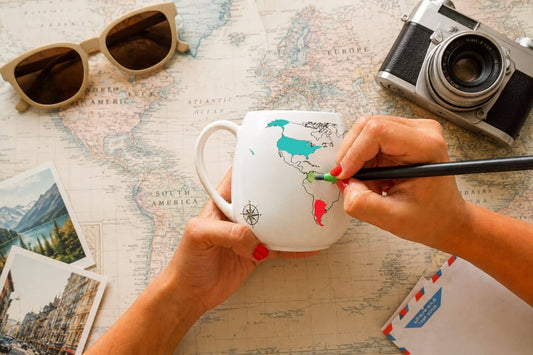 Hands colouring in a country on the world map colour-in mug using a green ceramic pen with travel props in the background.