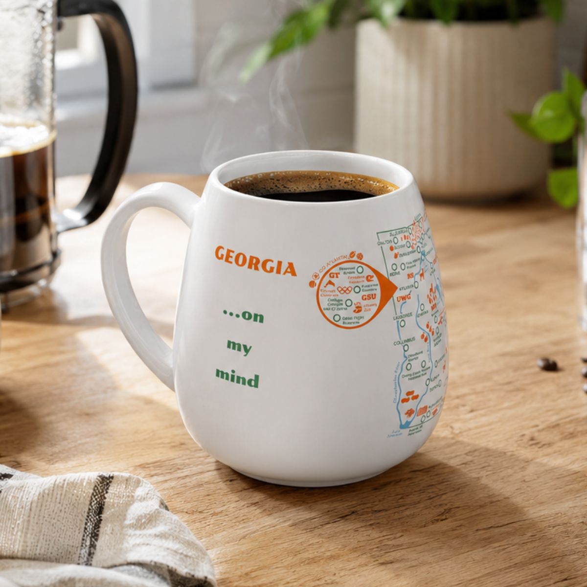 White mug with a Georgia map design to track trips and experiences placed on a wooden kitchen counter with coffee.