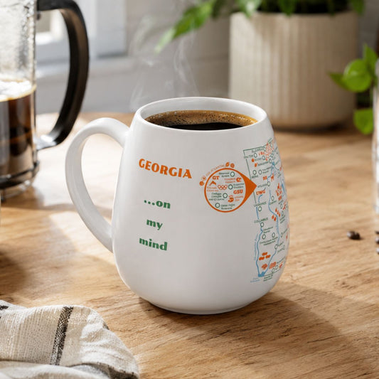 White mug with a Georgia map design to track trips and experiences placed on a wooden kitchen counter with coffee.