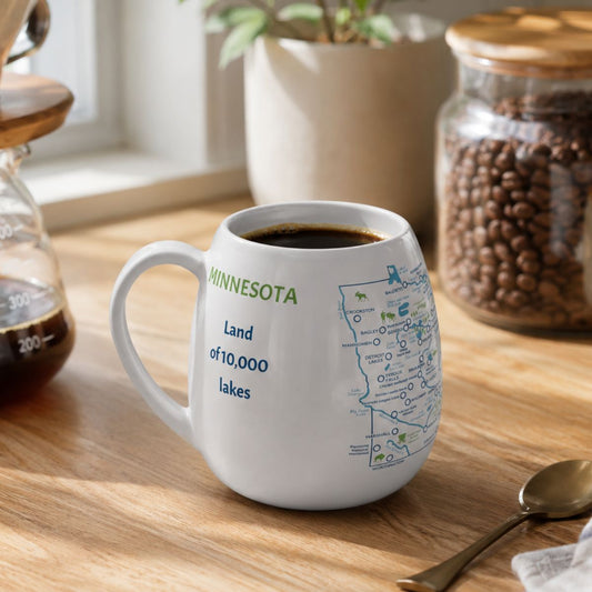 White mug with a Minnesota map design to track trips and experiences placed on a wooden kitchen counter with coffee.
