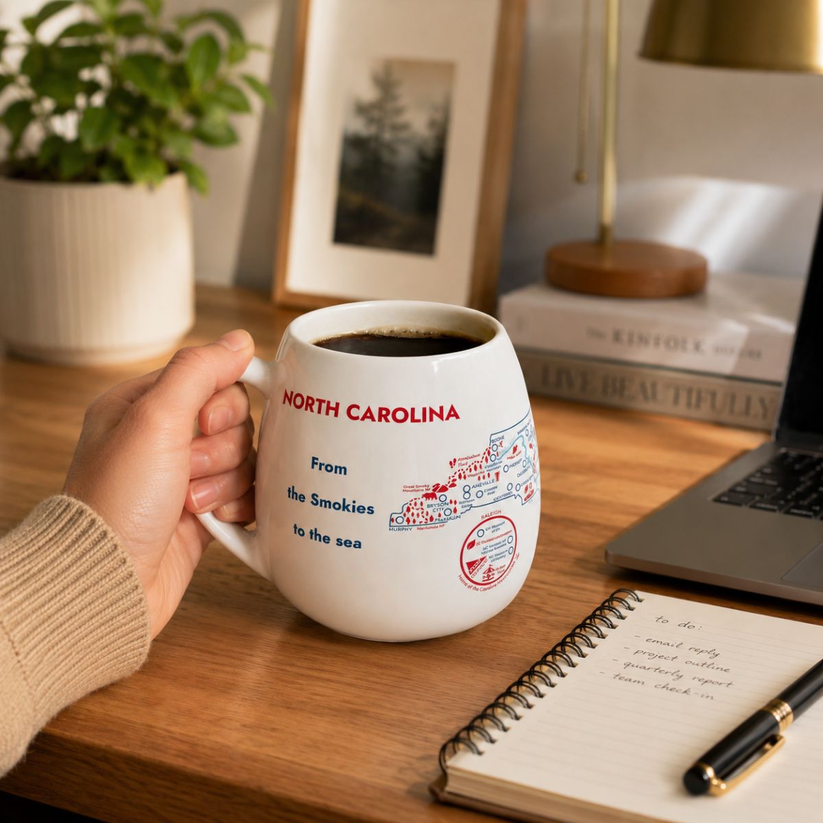 Person holding a North Carolina color-in mug on a desk with a laptop and notebook