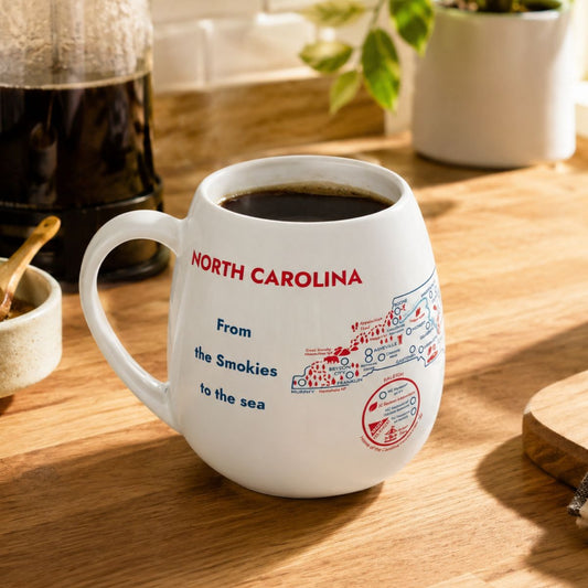 White mug with a North Carolina map design to track trips and experiences placed on a wooden kitchen counter with coffee.