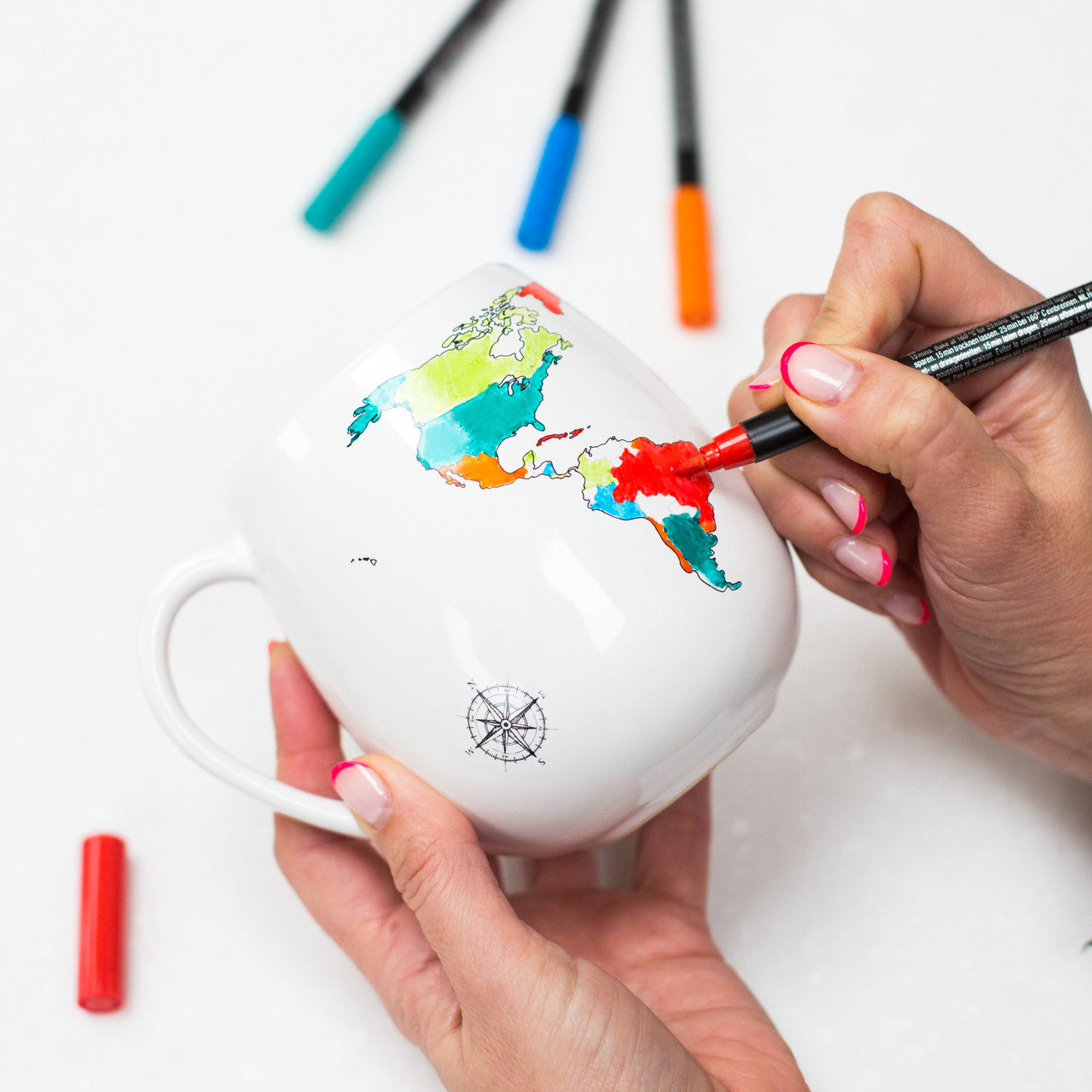 Hands holding and colouring in on the World Map Mug using a red ceramic pen with other ceramic pens in the background