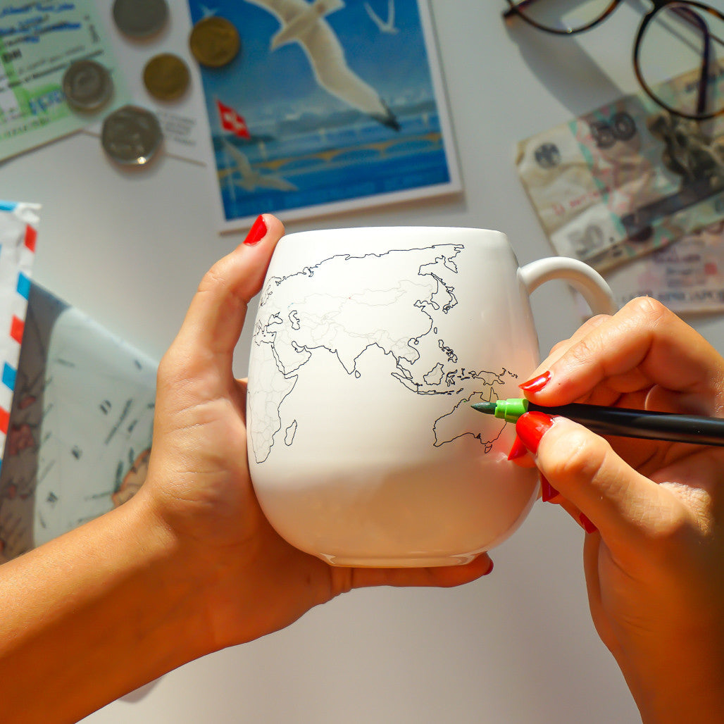 Hands colouring in countries on a world map mug with a green ceramic pen to track travel adventures, with coins and postcards in the background