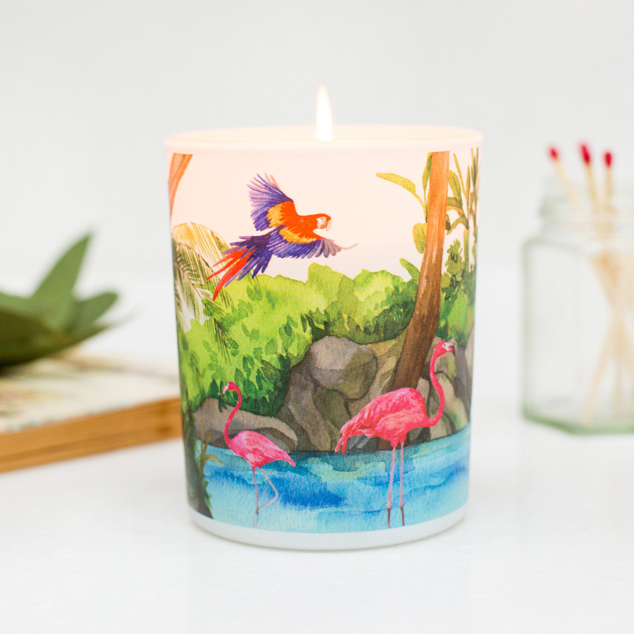 A colorful scented candle with a vibrant design featuring flamingos, macaws, and tropical plants on a glass jar, nature-inspired eco-friendly candle gift by Trouvaille