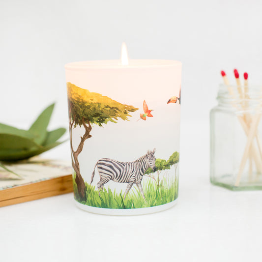Lit candle with a vibrant grassland design featuring a zebra, placed on a table with matches and a book in the background. A sustainable uplifting gift for nature lovers.