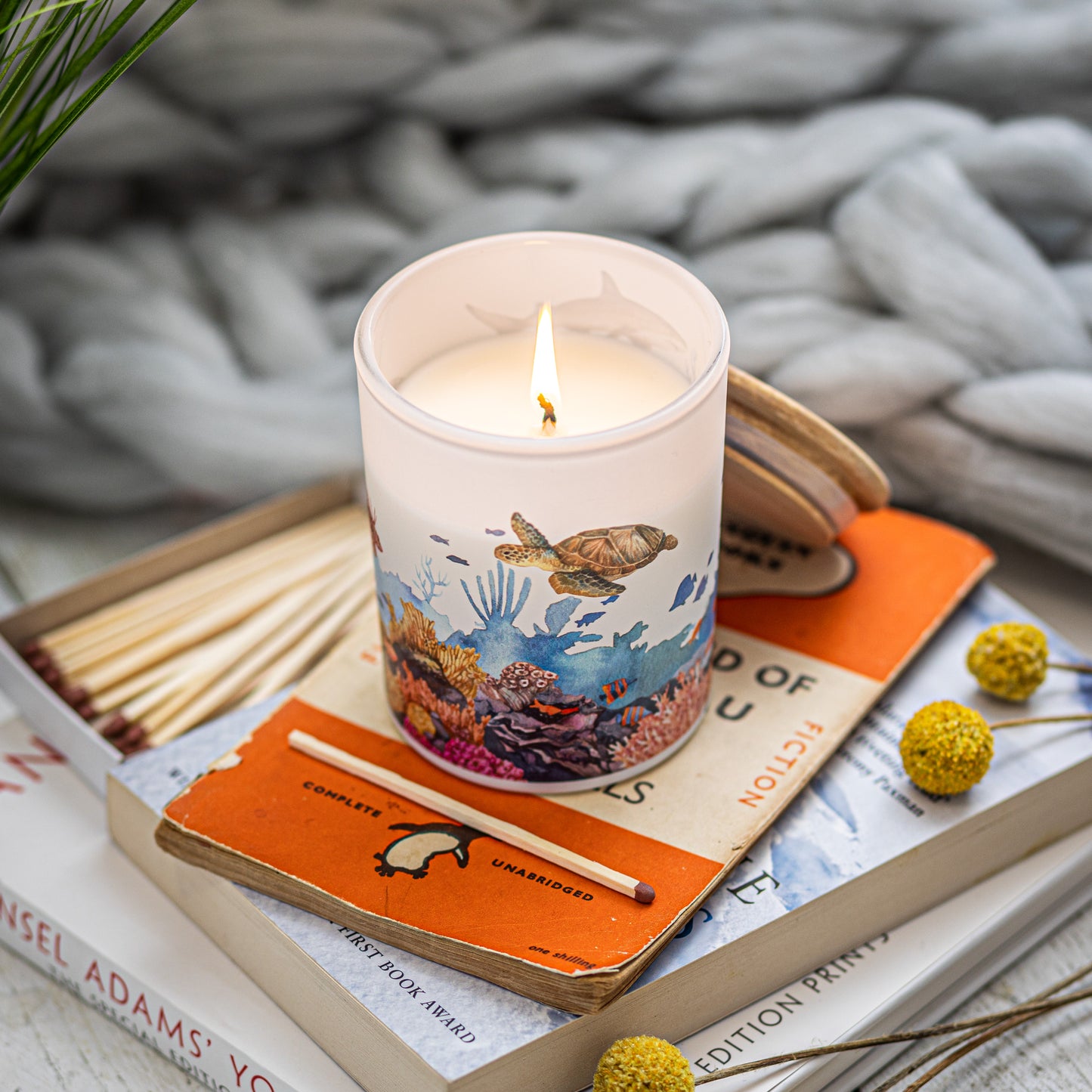 A lit candle with a marine life theme placed on an orange book, surrounded by yellow flowers and wooden matches, a sustainable home fragrance gift for ocean lovers.