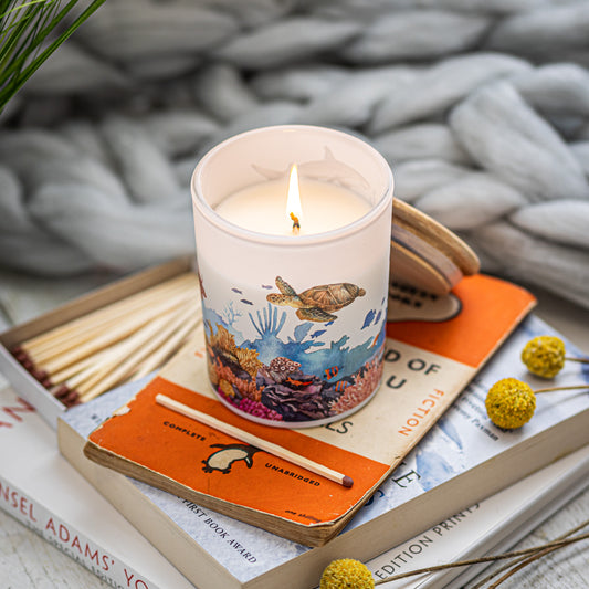 A lit candle with a marine life theme placed on an orange book, surrounded by yellow flowers and wooden matches, a sustainable home fragrance gift for ocean lovers.