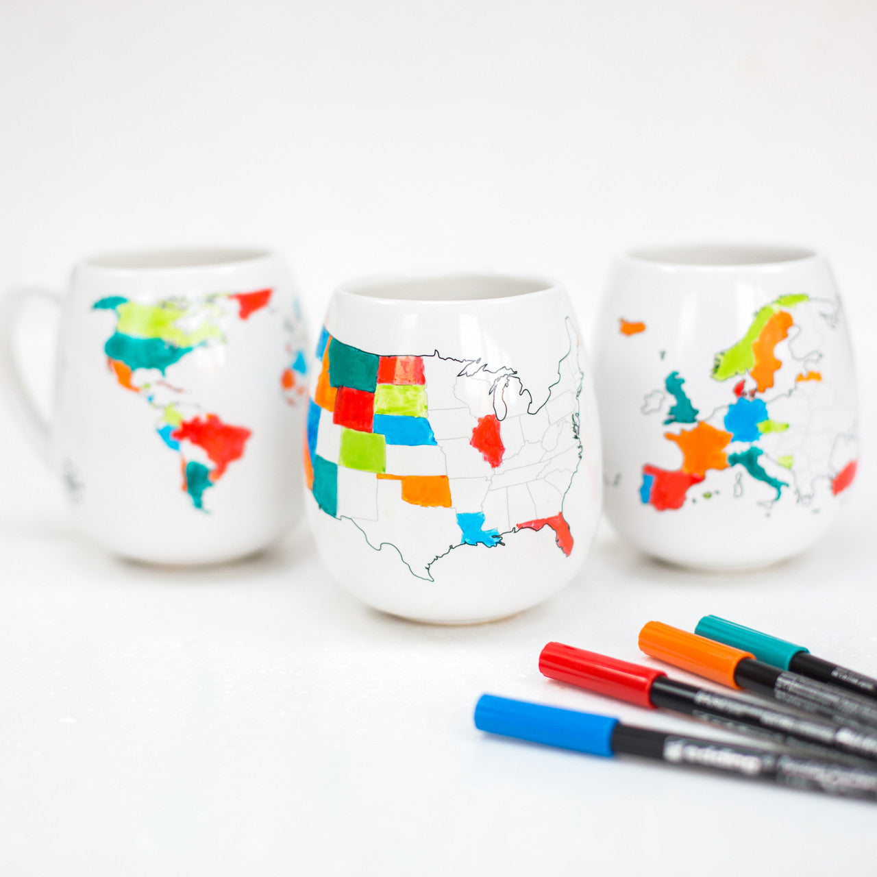 Coloured World mug, USA mug and Europe mug on a white background with four ceramic pens, including blue, orange, red and teal