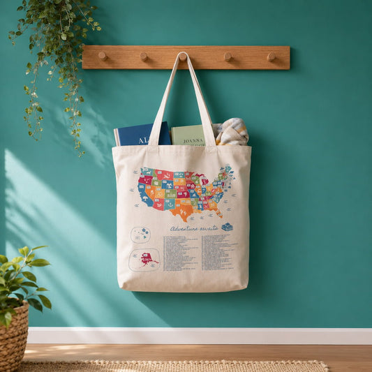 Lifestyle photo of the US Book Lover's Color In Totebag hanging from a wooden wall hook against a teal-green wall with natural sunlight and plants.
