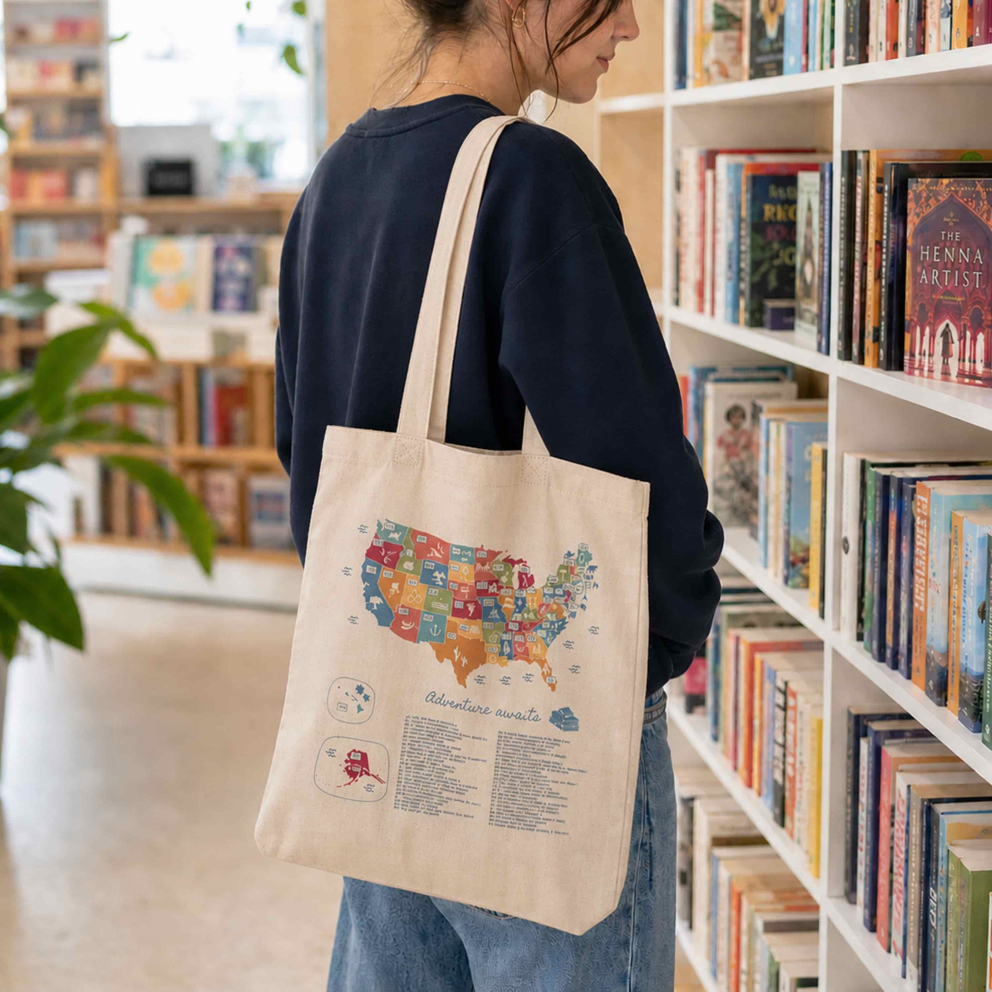 Person wearing a natural cotton tote bag with a USA map book tracker design in a bright modern bookstore, with shelves of books and plants in the background.
