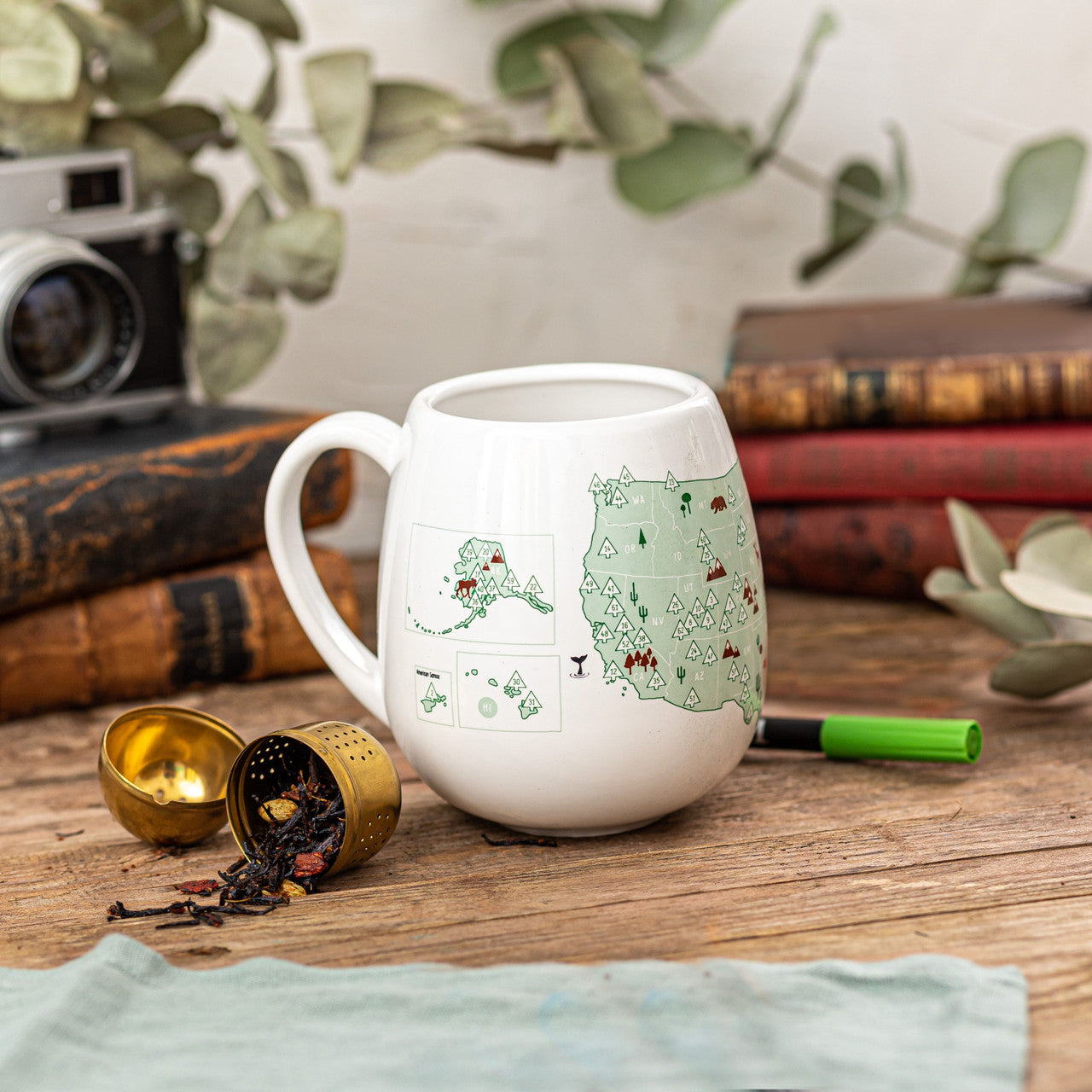 White ceramic mug with a map of the United States featuring trees representing different national parks, accompanied by a green ceramic pen on a wooden table.