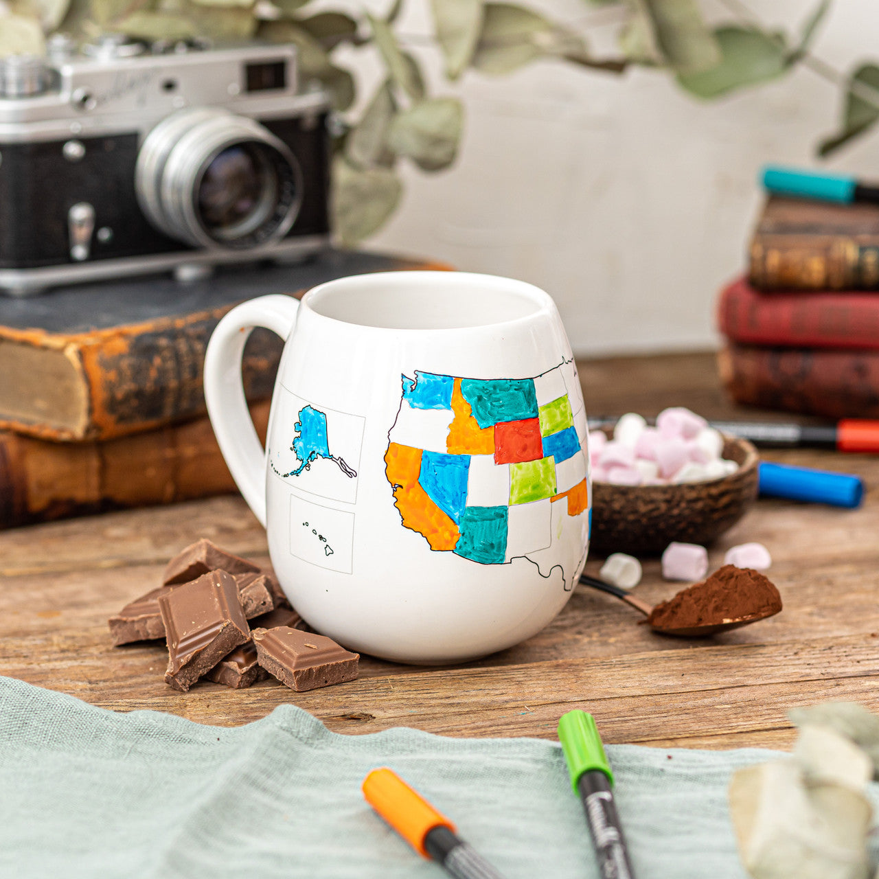 A white ceramic travel mug with a map of the United States with some states colored in, placed on a wooden surface with ceramic pens and chocolate pieces, unique gift for travelers