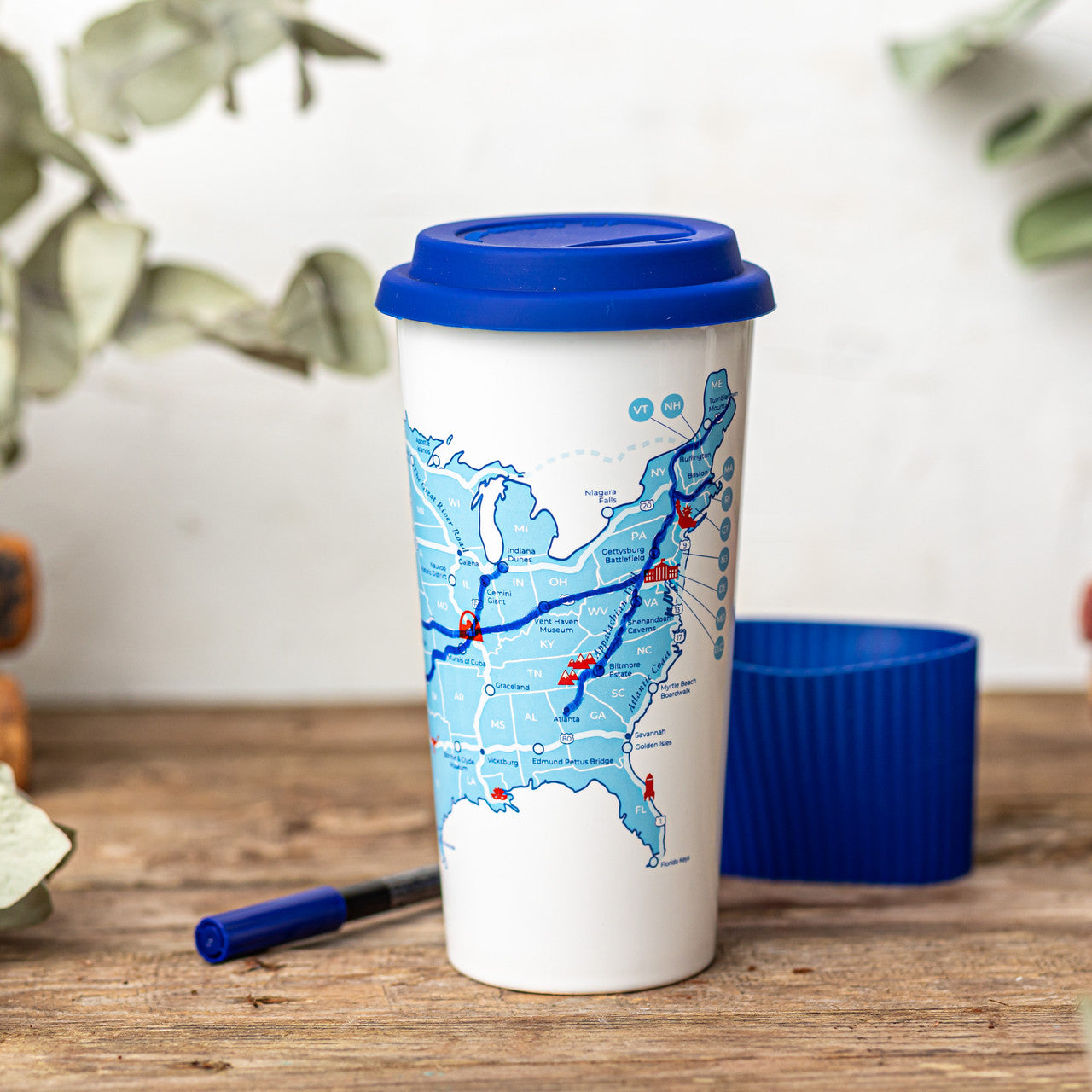 A white ceramic cup with a blue lid and sleeve featuring a colorful map of road trips in the United States to color in with the ceramic marker